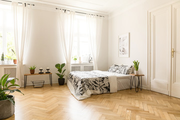 Patterned sheets on bed under poster in spacious bright bedroom interior with plants. Real photo - obrazy, fototapety, plakaty