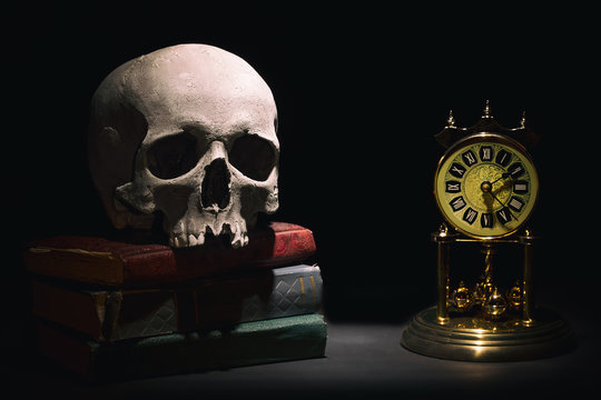 Human skull on old books near retro vintage clock on black background under beam of light. Dramatic concept.