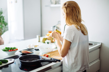 Back view of blonde housewife standing near the kitchen stove with frying pan, preparing meat...