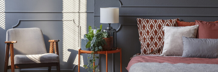 Decorative cushions on bed standing in dark grey room interior with molding on wall, metal bedside table and armchair