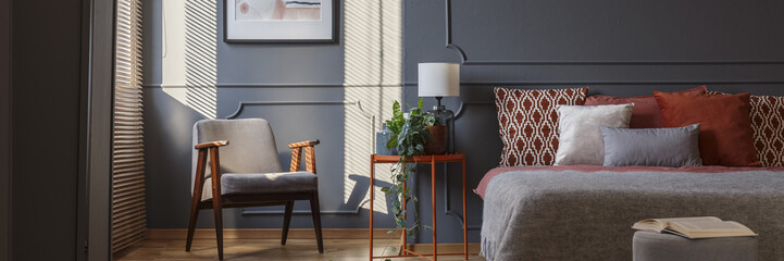 Grey armchair standing in dark grey bedroom interior with molding on the wall, orange beside table with plant and lamp and double bed with pillows - obrazy, fototapety, plakaty