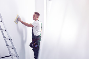 Smiling handyman cleaning white wall before painting while renovating interior - obrazy, fototapety, plakaty
