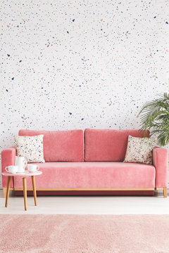 Pillows on pink sofa against wallpaper in living room interior with table and plant. Real photo