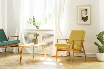 Poster above yellow wooden armchair at table in retro living room interior with window. Real photo