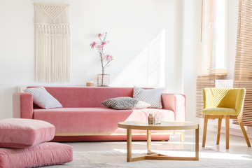 Real photo of a pink couch with pillows standing next to big pillows and yellow armchair, and behind a wooden table in bright living room interior with flowers and blinds on windows