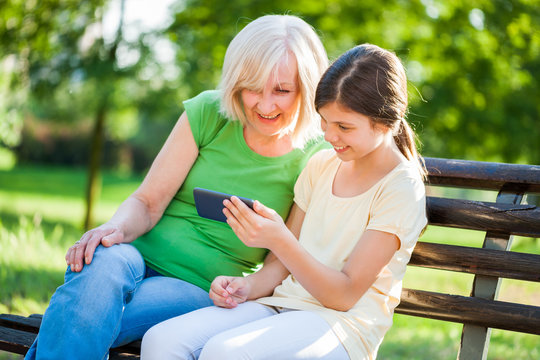 Grandmother and granddaughter are sitting in park and using mobile phone. 