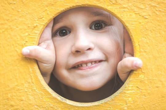 Face Of Small Child Girl Looking Through A Hole In A Play Equipment Outdoors. Yellow Background. Happy Childhood Concept.