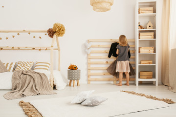Young girl hanging clothes on wooden hanger in white Nordic style bedroom interior with home-shape bed, two pillows placed on carpet and white rack with wooden boxes - obrazy, fototapety, plakaty