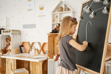 Little blonde girl drawing on the blackboard in her white room interior with wooden crate shelves, simple desk and posters on the wall