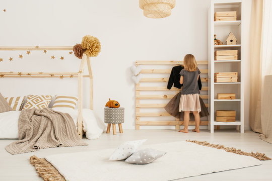 Young girl hanging clothes on wooden hanger in white Nordic style bedroom interior with home-shape bed, two pillows placed on carpet and white rack with wooden boxes