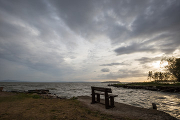 Fototapeta na wymiar A wooden sitting bench on a lake shore at sunset, beneath a moody, cloudy sky