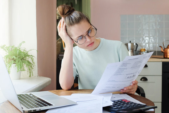 Young frustrated unhappy tired woman with financial troubles, sitting at kitchen table with papers, calculator and laptop computer, reading documents.