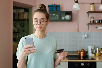 Closeup shot of young smiley woman wearing trendy round eyeglasses, paying with credit card while...