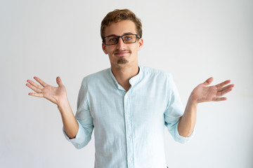 Smiling careless blonde guy showing do not know gesture. Young Caucasian man in glasses throwing up...