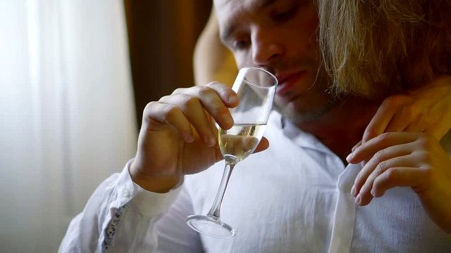 young woman is seducing adult man, sitting in a chair with glass of champagne in a hand, standing behind him