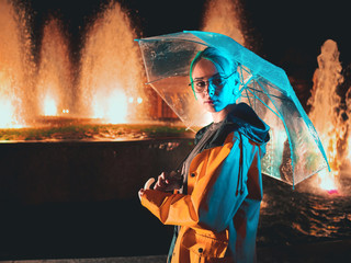 Young pretty girl with blue dyed hair in yellow raincoat and with transparent umbrella stands near fountain. Night neon illumination of city. Portrait of stylish hipster with glasses.