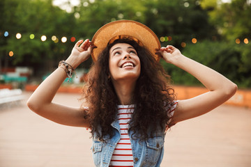 Photo of pretty young brunette woman 18-20 wearing straw hat, enjoying summer and walking in green...