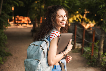 Photo of caucasian young brunette woman 18-20 wearing backpack, walking through green park with silver laptop in hands