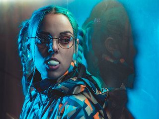 Young pretty girl with unusual hairstyle inflates the chewing bubble near glowing turquoise neon...