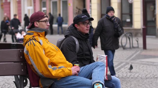 Friends teenagers sit on bench eat chips and drink cola from a can in public city center place