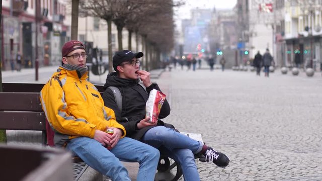Friends teenagers sit on bench eat chips and drink cola from a can in public city center place