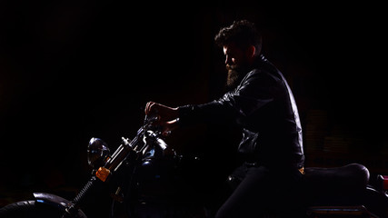 Night rider concept. Man with beard, biker in leather jacket sitting on motor bike in darkness, black background. Hipster, brutal biker in leather jacket riding motorcycle at night time, copy space.