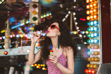 Young pretty lady in heart sunglasses and dress standing and blowing bubbles while spending time in amusement park