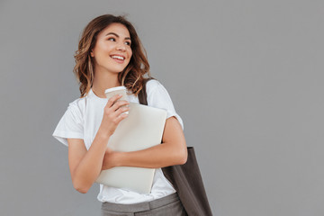 Image of attractive brunette woman 20s smiling and looking aside at copyspace while standing against gray wall isolated with silver laptop, and takeaway coffee in hands