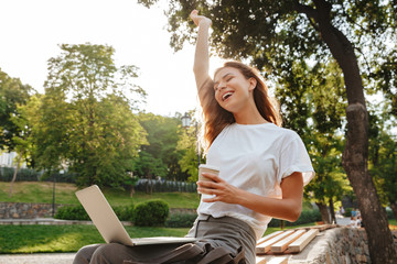 Image of happy modern woman sitting on bench in green park and enjoying summertime, while using silver laptop and drinking takeaway coffee from paper cup