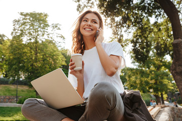 Image of smiling modern woman sitting on bench in green park on summer day, and talking on smartphone while using silver laptop and drinking takeaway coffee