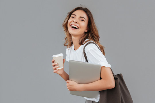 Image of happy young woman 20s laughing and standing over gray wall isolated with silver laptop, and takeaway coffee in hands
