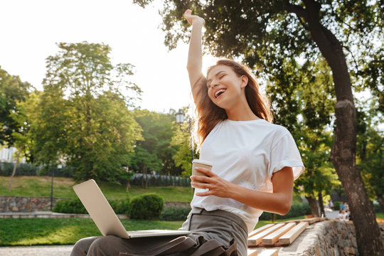 Image of happy modern woman sitting on bench in green park and enjoying summertime, while using silver laptop and drinking takeaway coffee from paper cup