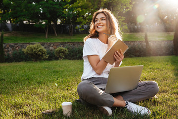 Portrait of beautiful smiling woman sitting on green grass in park with legs crossed during summer...