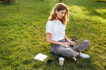 Photo of european woman 20s sitting on green grass in park with legs crossed during summer day,...