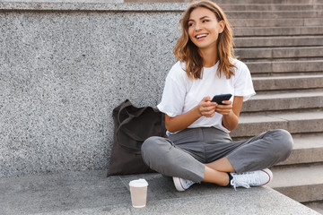 Image of beautiful stylish woman sitting on street stairs with legs crossed on summer day, and holding mobile phone - Powered by Adobe