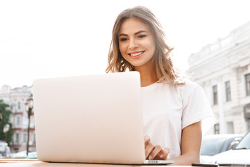 Photo of pretty young woman sitting at table outdoor on summer day, and using silver laptop