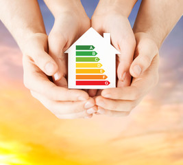 efficiency and home concept - close up of female hands holding paper house with energy saving rating over evening sky background