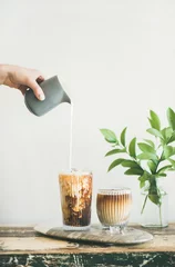  Iced coffee in tall glasses with milk poured over from pitcher by hand, white wall and green plant branches in vase at background, copy space. Summer refreshing beverage © sonyakamoz
