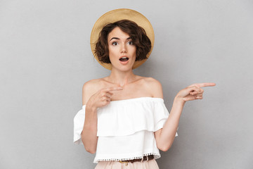 Beautiful brunette woman 20s wearing straw hat looking at you and pointing finger at copyspace with open mouth, isolated over gray background