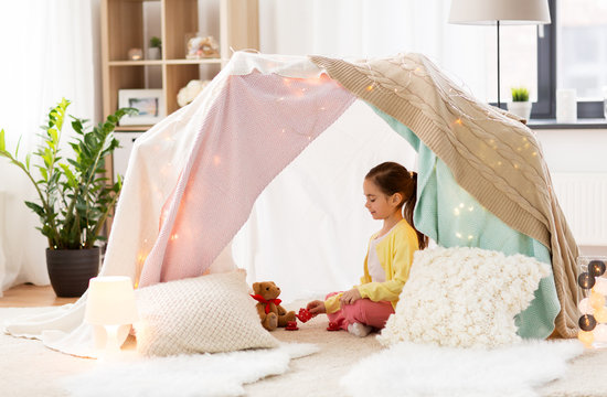childhood and hygge concept - happy little girl playing tea party with teddy bear in kids tent at home
