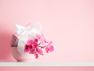 Gift box with white satin ribbon and pink orchid on a white shelf and on the background of a pink wall. Greeting card for Mother's Day, Easter, Happy Woman's Day, Wedding, Birthday, Valentine's Day 