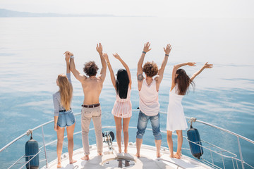 Group of caucasian guys and their girlfriends standing with raised hands on the elge of yacht s deck, happy with sun, sea wind and holyday time. The concept of happiness and freedom.