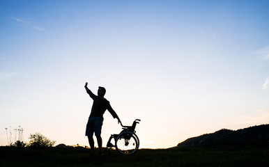 A young man standing by wheelchair in nature in the evening.