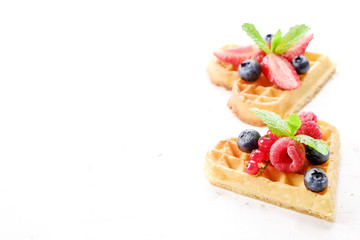 Heart shape belgium waffles with assorted berries mix, strawberry, blueberry, raspberry and red currant decorated with mint leaf on white ceramic plate. Close up, copy space, background, top view.