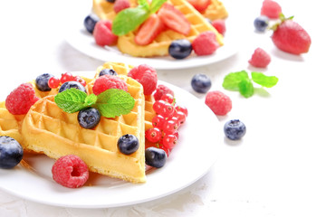 Heart shape belgium waffles with assorted berries mix, strawberry, blueberry, raspberry and red currant decorated with mint leaf on white ceramic plate. Close up, copy space, background, top view.