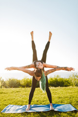 Young ladies in black sporty tops and leggings training yoga poses outdoors. Pretty woman holding on her back girl while practicing yoga together