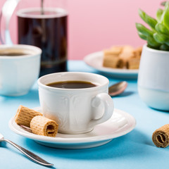 Beautiful composition with two cups of coffee with crispy mini wafer rolls and indoor plants succulent on blue and pink background.