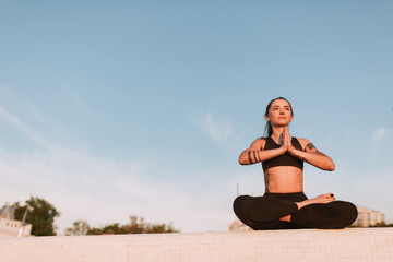 Fototapeta na wymiar Beautiful lady sitting in lotus pose and dreamily looking aside. Young woman in black sporty top and leggings meditating while practicing yoga