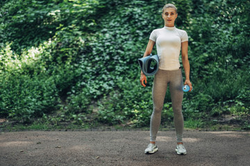 Fitness Woman Preparing for Yoga in the Early Morning Park. Wearing Stylish Sport Outfit. Blue Bottle of Water and Yoga Mat. Healthy Lifestyle.