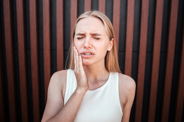 Beautiful blonde teen is standing and holding hand on cheeck. She has a tooth pain. Girl is upset....